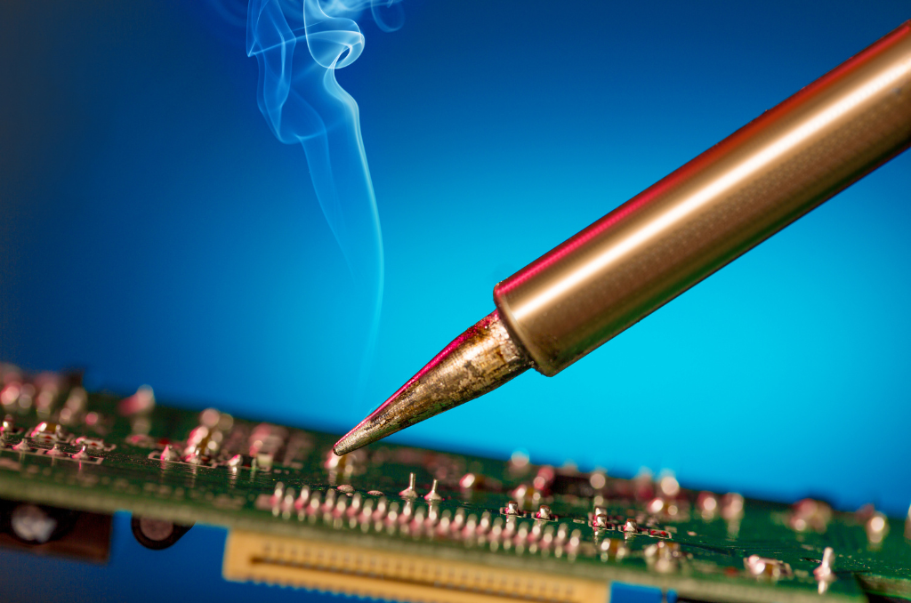 Close up side image of a small thin soldering iron working on a green circuit board with a blue background