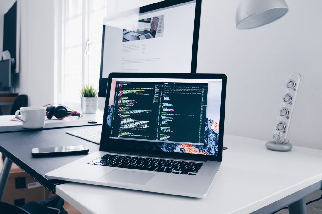 Laptop programming in Python on a white desk with a monitor and window in the background