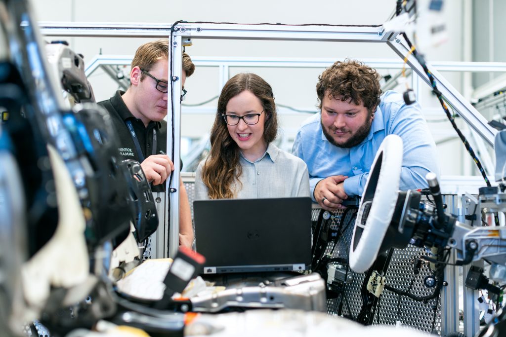 An engineering team, two men and one woman, looking at a laptop while surrounded by automotive equipment
