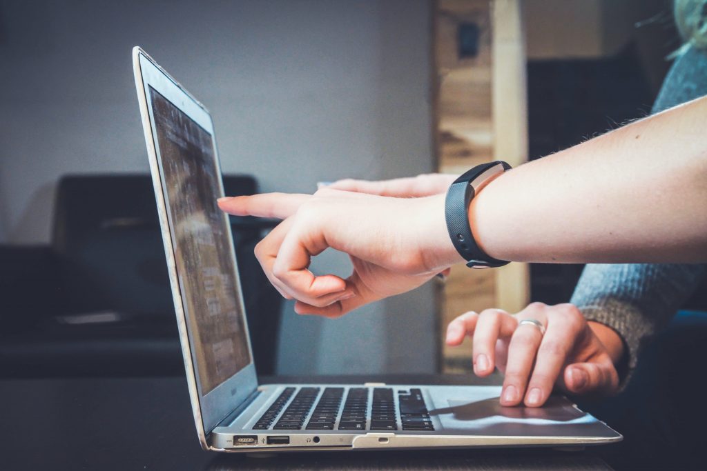 Closeup sideview of laptop. A hand that is wearing a watch is pointing to the screen while another hand uses the touchpad