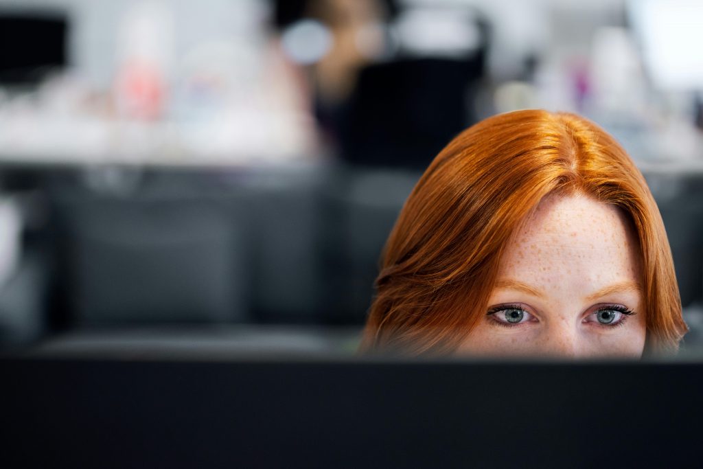 Closeup of the top of a redheaded woman's head behind a computer monitor